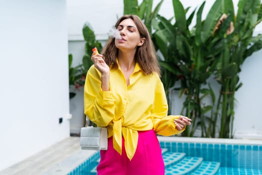 Stylish fit fashion women in bright pink pants holding bag trendy posing at luxury tropical villa by pool outdoor natural day light smoking vape pod enjoying puffs