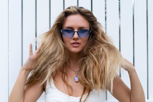 Beautiful young blonde woman in a top with long hair and smooth clean soft skin, natural beauty, daylight, wearing blue glasses, posing against the backdrop of a wooden fence