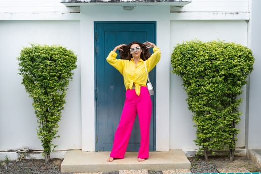 Stylish fit fashion women in bright pink wide leg pants and yellow shirt holding bag trendy mint sunglasses posing at luxury tropical villa by pool outdoor natural day light