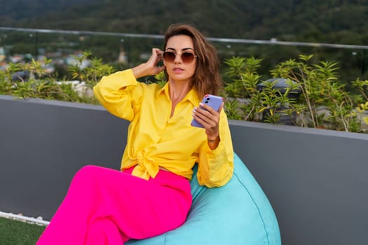 Stylish fit fashion women in bright pink wide leg pants and yellow shirt holding bag trendy sunglasses posing at rooftop terrace tropical view outdoor holding mobile phone