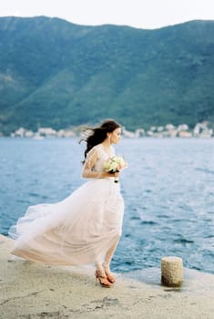 Bride in a dress fluttering in the wind stands on the pier and looks at the sea. High quality photo
