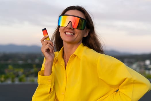 Stylish fit fashion women in bright yellow shirt trendy shield visor rainbow sunglasses posing at rooftop terrace tropical view sunset smoking vape pod enjoying puffs