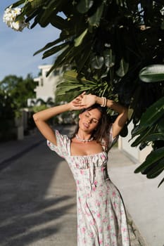 Young beautiful woman in a romantic dress with a floral print, and a pearl necklace bracelet, against the backdrop of tropical leaves