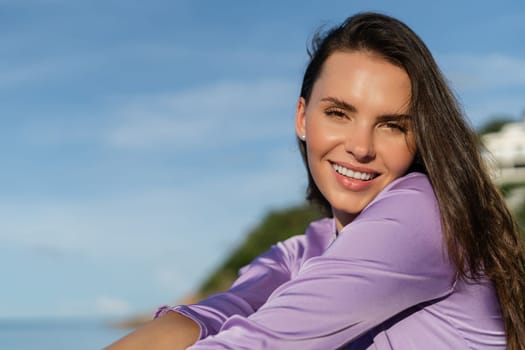 A young beautiful woman in a romantic mood, lilac silk clothes, on the beach against the backdrop of the sea and stones at sunset, laughs, smiles with a snow-white smile