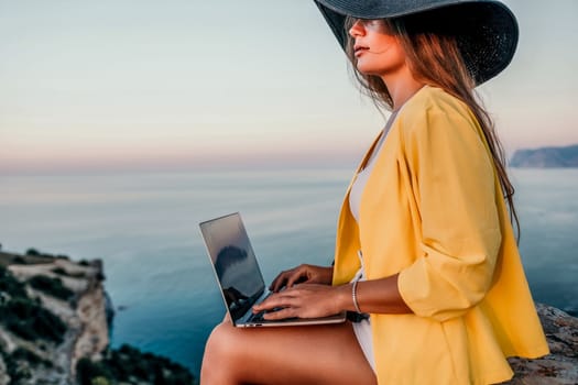 Successful business woman in yellow hat working on laptop by the sea. Pretty lady typing on computer at summer day outdoors. Freelance, travel and holidays concept.
