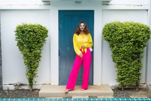 Stylish fit fashion women in bright pink wide leg pants and yellow shirt holding bag trendy mint sunglasses posing at luxury tropical villa by pool outdoor natural day light