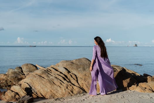 Young beautiful woman in a romantic mood, lilac silk clothes, on the beach against the backdrop of the sea and stones at sunset