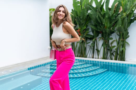 Stylish fit fashion women in bright pink wide leg pants holding bag trendy mint sunglasses posing at luxury tropical villa by pool outdoor natural day light