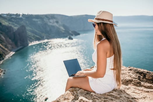 Successful business woman in yellow hat working on laptop by the sea. Pretty lady typing on computer at summer day outdoors. Freelance, travel and holidays concept.