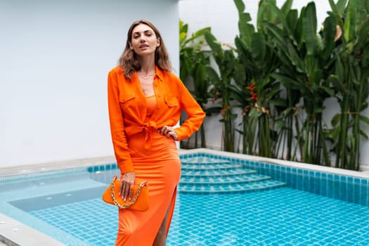 Stylish fit fashion women in bright orange fitting dress and shirt holding fashion bag posing at luxury tropical villa by pool outdoor natural day light