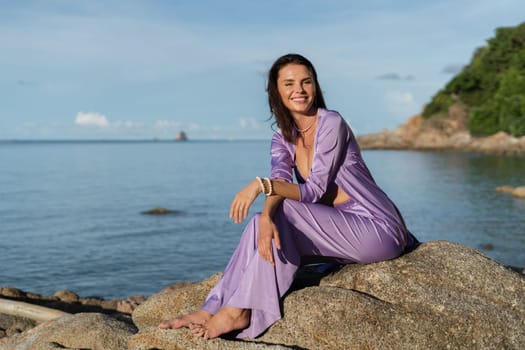 A young beautiful woman in a romantic mood, lilac silk clothes, on the beach against the backdrop of the sea and stones at sunset, laughs, smiles with a snow-white smile