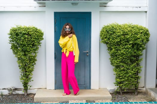 Stylish fit fashion women in bright pink wide leg pants and yellow shirt holding bag trendy mint sunglasses posing at luxury tropical villa by pool outdoor natural day light