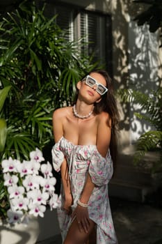 Young beautiful woman in a romantic dress with a floral print, sunglasses and a pearl necklace bracelet, against the backdrop of tropical leaves