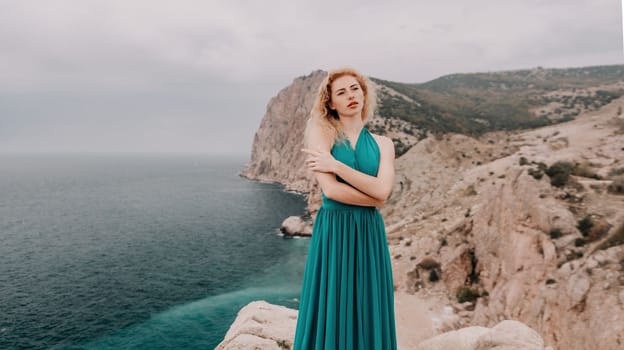 Side view a Young beautiful sensual woman in a mint long dress posing on a volcanic rock high above the sea during sunset. Girl on the nature on overcast sky background. Fashion photo
