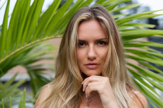 Beautiful young blonde in a top with long hair and smooth clean soft skin, natural beauty, daylight, pendant on neck accessory, posing with palm leaf