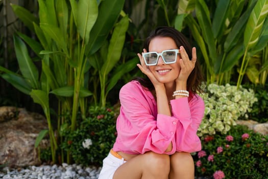 Young beautiful brunette in a pink shirt, neck jewelry, necklace, trendy sunglasses sits on a background of tropical leaves, laughs, smiles