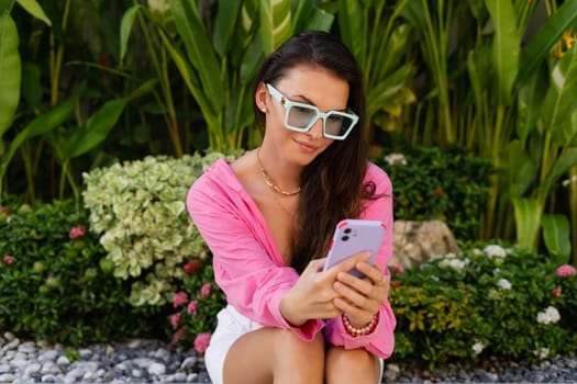 A young beautiful brunette woman in a pink shirt, jewelry on her neck, trendy sunglasses sits on a background of tropical leaves, looks at the phone screen, smiles