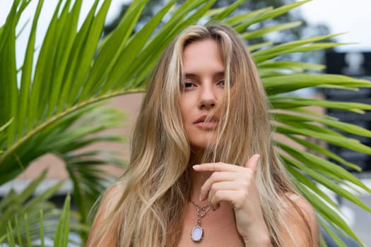 Beautiful young blonde in a top with long hair and smooth clean soft skin, natural beauty, daylight, pendant on neck accessory, posing with palm leaf