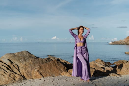 Young beautiful woman in a romantic mood, lilac silk clothes, on the beach against the backdrop of the sea and stones at sunset