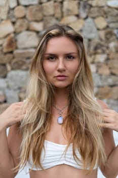 Beautiful young blonde in a top with long hair and smooth clean soft skin, natural beauty, daylight, pendant on neck accessory, posing on brick wall