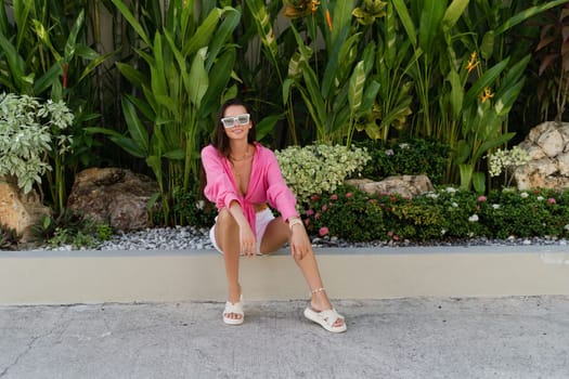 Young beautiful brunette in a pink shirt, neck jewelry, necklace, trendy sunglasses sits on a background of tropical leaves, laughs, smiles