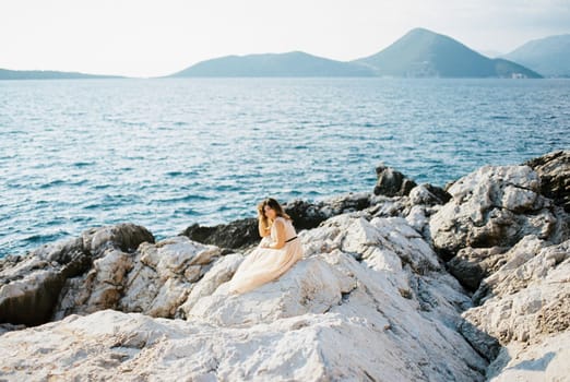 Bride sits on a rocky seashore against the backdrop of mountains, propping her head on her hand. High quality photo