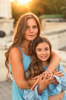 Portrait of mother and daughter in blue dresses with flowing long hair against the backdrop of sunset. The woman hugs and presses the girl to her. They are looking at the camera