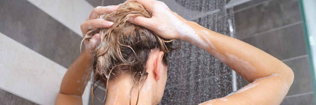 Young woman washes her hair with shampoo and hands with foam closeup. Back of beautiful woman taking shower and enjoying body and hair hygiene