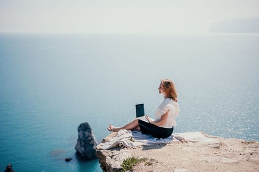 Successful business woman in yellow hat working on laptop by the sea. Pretty lady typing on computer at summer day outdoors. Freelance, travel and holidays concept.