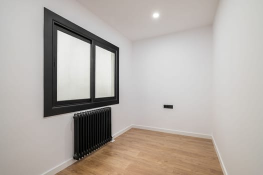 An empty room with a modern version of a plastic window with a black frame and opaque glass. Radiator for heating black. Bright lighting from the ceiling and wooden parquet