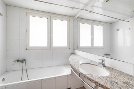 White bathroom with large hot tub. On the wall are two plastic windows with frosted glass through which daylight shines. Nearby is a sink on a marble countertop with a large mirror on the wall