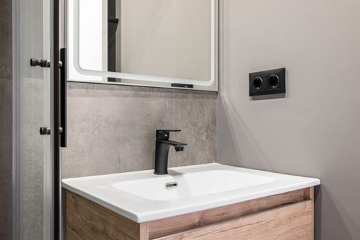 Closeup of a white square ceramic sink on a wooden vanity. Fittings and faucet made of ferrous metal. On the wall is a designer mirror with a white frame. Walls in gray pleasant tones
