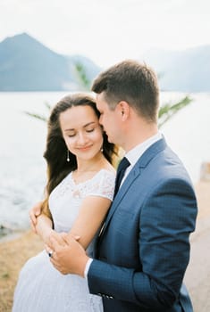 Groom hugs the bride, who closed her eyes by the shoulders on the seashore. Portrait. High quality photo