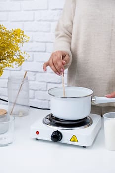 Creative hobby at home. Woman making decorative aroma candle at table, stirring melting soy wax in the pot