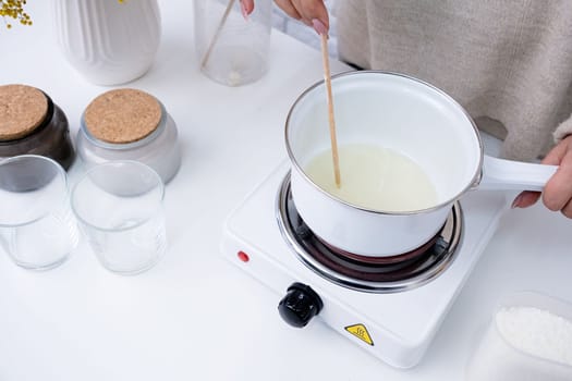 Creative hobby at home. Woman making decorative aroma candle at table, stirring melting soy wax in the pot
