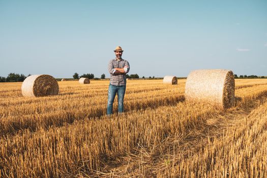 Happy farmer is standing beside bales of hay. He is satisfied because of successful harvesting.