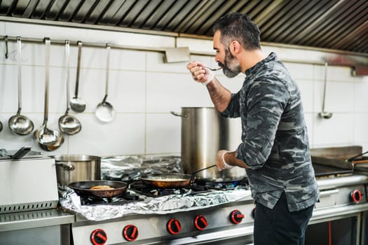 Professional cook is preparing meal in restaurant's kitchen. He is tasting meal.