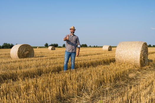 Happy farmer is standing beside bales of hay. He is satisfied because of successful harvesting.