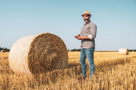 Happy farmer is standing beside bales of hay. He is examining straw after successful harvesting.