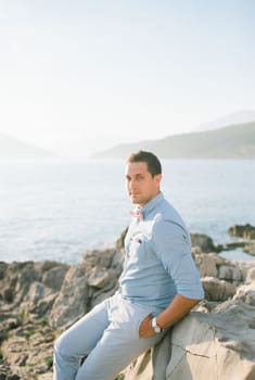 Groom sits on a rocky shore by the sea holding his hands in his pockets. High quality photo