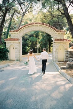 Bride and groom walk holding hands to the arched entrance to the park. High quality photo