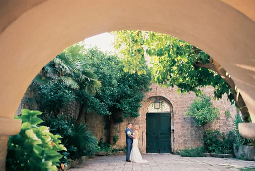 View from the arch to the hugging bride and groom in the courtyard of an ancient building. High quality photo