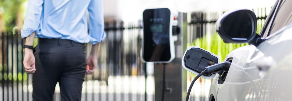 Closeup progressive suit-clad businessman with his electric vehicle recharge his car on public charging station in modern city with power cable plug and renewable energy-powered electric vehicle.