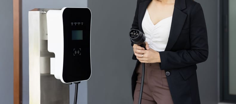 Closeup asian woman holding and pointing an EV plug, a home charging station providing a sustainable power source for electric vehicles. Alternative energy for progressive lifestyle.