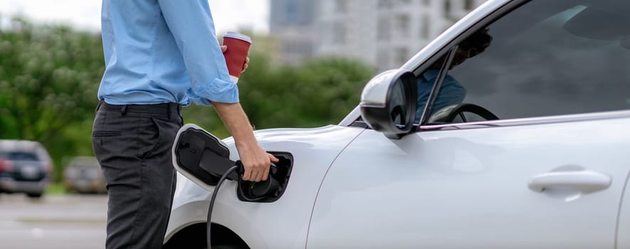 Progressive eco-friendly concept of parking EV car at public electric-powered charging station in city with blur background of businessman leaning on recharging-electric vehicle with coffee.