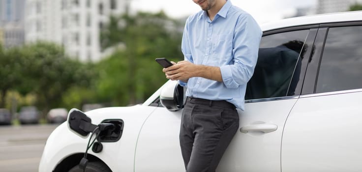 Progressive businessman talking on the phone, leaning on electric car recharging with public EV charging station, apartment condo residential building on the background as green city lifestyle.