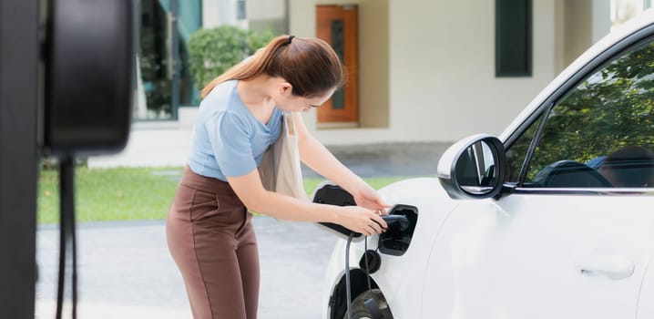 Progressive woman install cable plug to her electric car with home charging station. Concept of the use of electric vehicles in a progressive lifestyle contributes to clean environment.