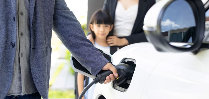 Focus closeup hand charging electric car, insert charger device into electric vehicle as progressive lifestyle concept of alternative green energy technology with blurred family in the background.