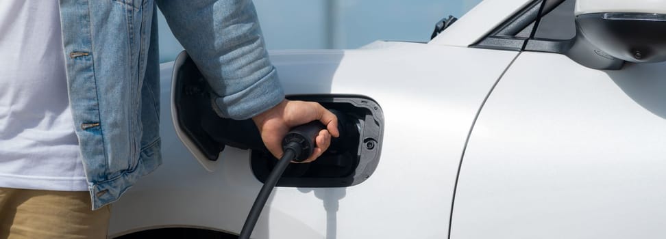 Progressive man with his electric car, EV car recharging energy from charging station on green field with wind turbine as concept of future sustainable energy. Electric vehicle with energy generator.