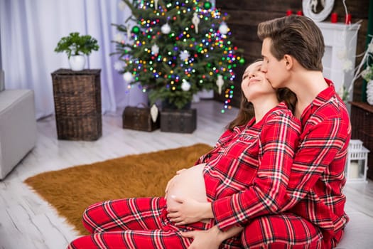 A pregnant woman has her hands on her stomach. A husband embraces his wife. The man kisses his partner on the cheek. A Christmas tree, a potted plant and a brown carpet can be seen in the background.